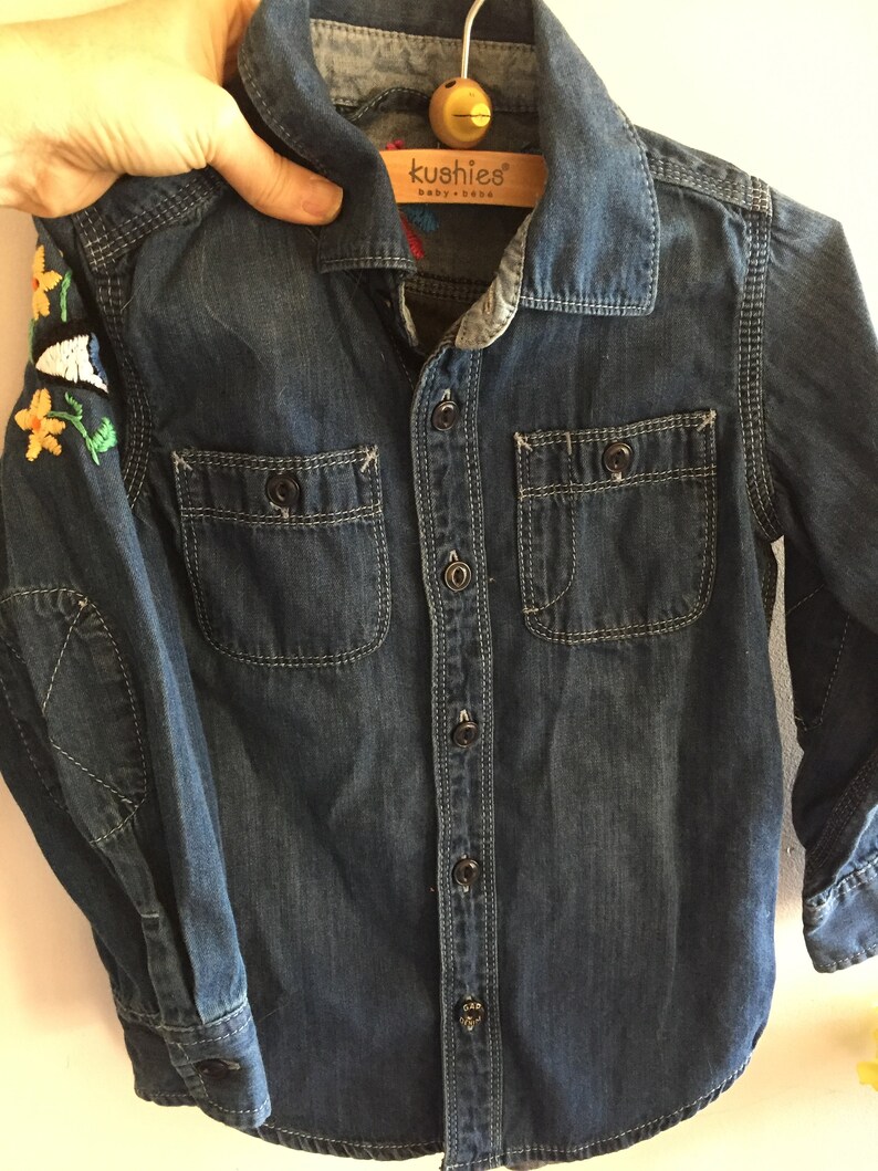 May include: A dark blue denim shirt with long sleeves and a button-up front. The shirt has two chest pockets and embroidered flowers on the left sleeve. The shirt has a label that reads "kushies baby + baba".