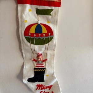 May include: A white felt Christmas stocking with a red cuff and a green, yellow, and blue hot air balloon with a small elf doll hanging from it. The stocking has the words "Merry Christmas" embroidered on it.
