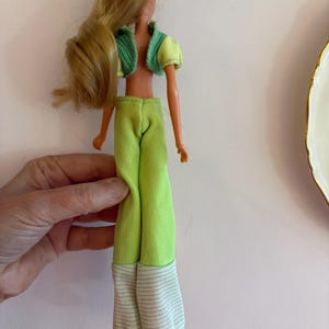 May include: A vintage doll with long blonde hair, dressed in a lime green outfit. The outfit features a cropped jacket with a green collar, matching trousers, and striped cuffs. The doll is wearing brown shoes.