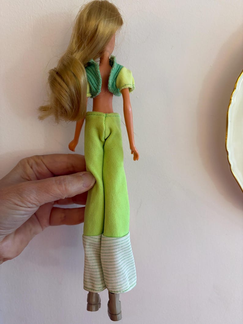 May include: A vintage doll with long blonde hair, dressed in a lime green outfit. The outfit features a cropped jacket with a green collar, matching trousers, and striped cuffs. The doll is wearing brown shoes.