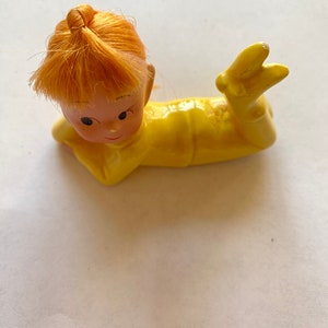 May include: A yellow ceramic figurine of a child with orange hair lying on its side. The child is wearing a yellow jumpsuit and has a painted face.
