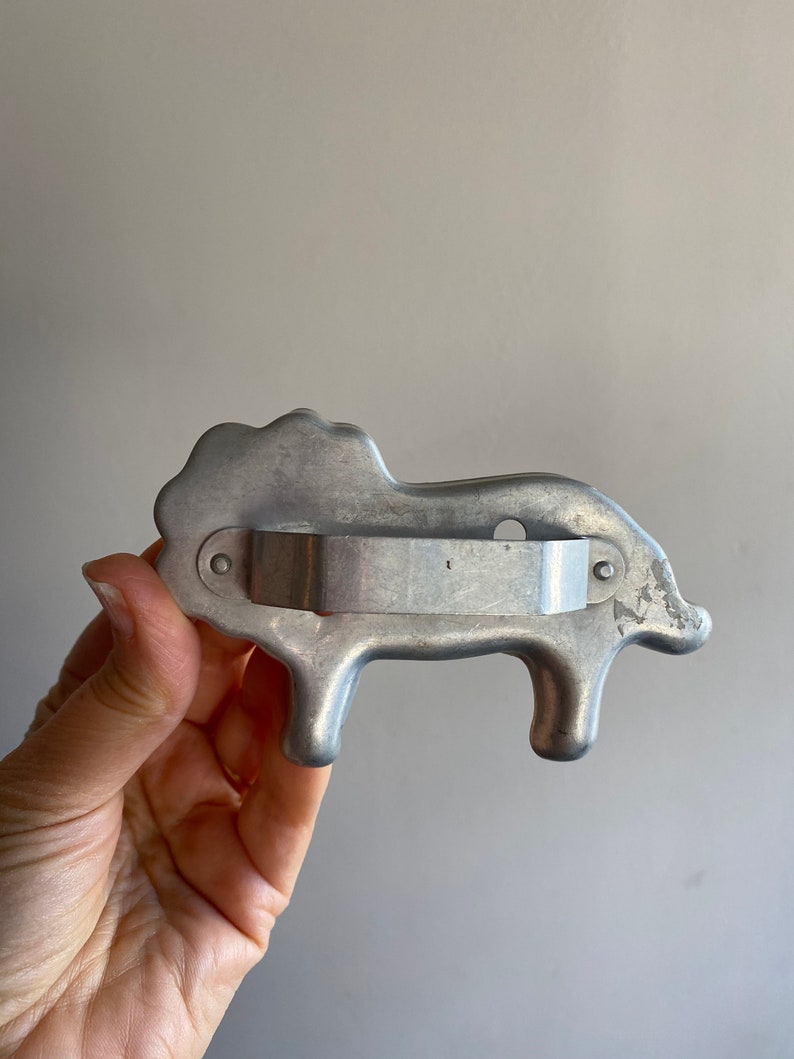 Puede incluir: Un cortador de galletas de metal plateado con forma de cerdo. El cortador tiene un mango en la parte posterior.