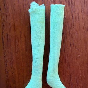 May include: A pair of pale green knee-high socks. One sock features a decorative pattern. The socks are made of a ribbed knit fabric. The socks are displayed on a brown wooden surface.