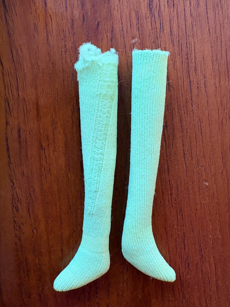 May include: A pair of pale green knee-high socks. One sock features a decorative pattern. The socks are made of a ribbed knit fabric. The socks are displayed on a brown wooden surface.
