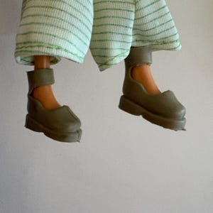 May include: A doll's legs and feet are shown, wearing olive green shoes with ankle straps. The doll is also wearing light green and white striped trousers. The image is set against a plain white background.