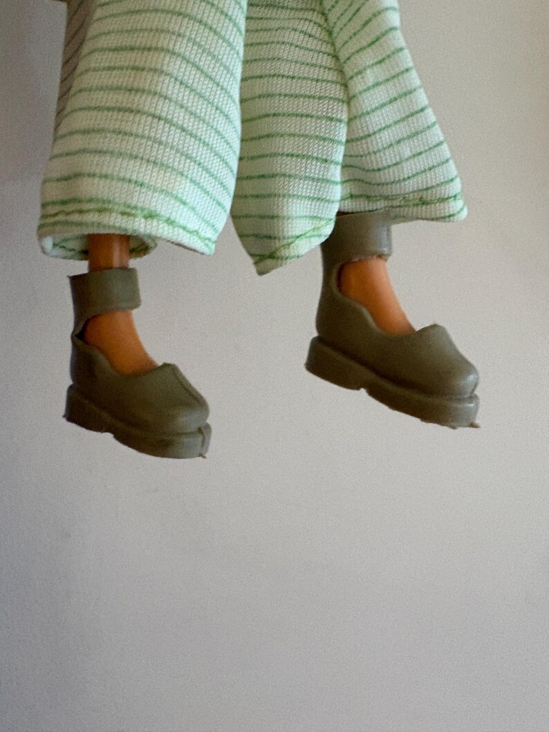 May include: A doll's legs and feet are shown, wearing olive green shoes with ankle straps. The doll is also wearing light green and white striped trousers. The image is set against a plain white background.