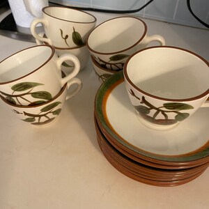 May include: Set of six white teacups and saucers with a brown rim and green leaf design. The saucers are stacked on top of each other.