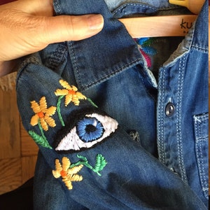 May include: A blue denim shirt with embroidered yellow flowers and a blue eye on the sleeve. The shirt has a button-up closure and a collar.