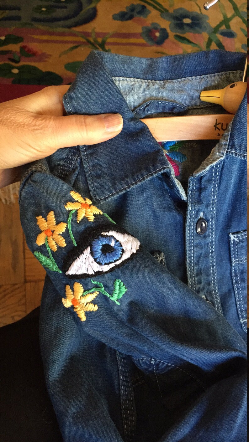 May include: A blue denim shirt with embroidered yellow flowers and a blue eye on the sleeve. The shirt has a button-up closure and a collar.