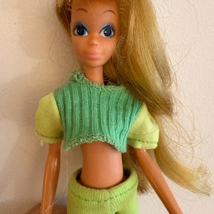 May include: A vintage doll with long blonde hair and blue eyes. The doll is wearing a green cropped sweater and matching trousers. The doll's arms and legs are visible, and the doll is posed against a plain background.