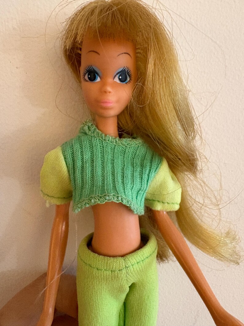 May include: A vintage doll with long blonde hair and blue eyes. The doll is wearing a green cropped sweater and matching trousers. The doll's arms and legs are visible, and the doll is posed against a plain background.