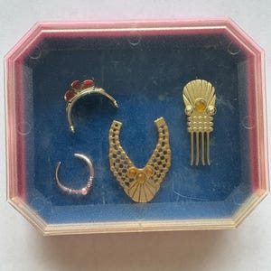Peut inclure: Un ensemble de petits bijoux dor&eacute;s pr&eacute;sent&eacute;s dans un r&eacute;cipient en plastique transparent &agrave; bord rose. L'ensemble comprend un diad&egrave;me, un collier, un peigne &agrave; cheveux et une petite boucle d'oreille. Le fond est bleu fonc&eacute;.