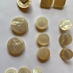 May include: A collection of 13 white mother of pearl buttons in various shapes and sizes. The buttons have a smooth, glossy finish and some have a rope-like design around the edge.