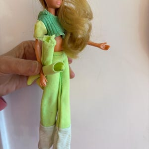 May include: A vintage doll with blonde hair and blue eyes, dressed in a green and yellow crop top, green trousers, and brown shoes. The trousers have white and grey striped cuffs. The doll is a collectible item.
