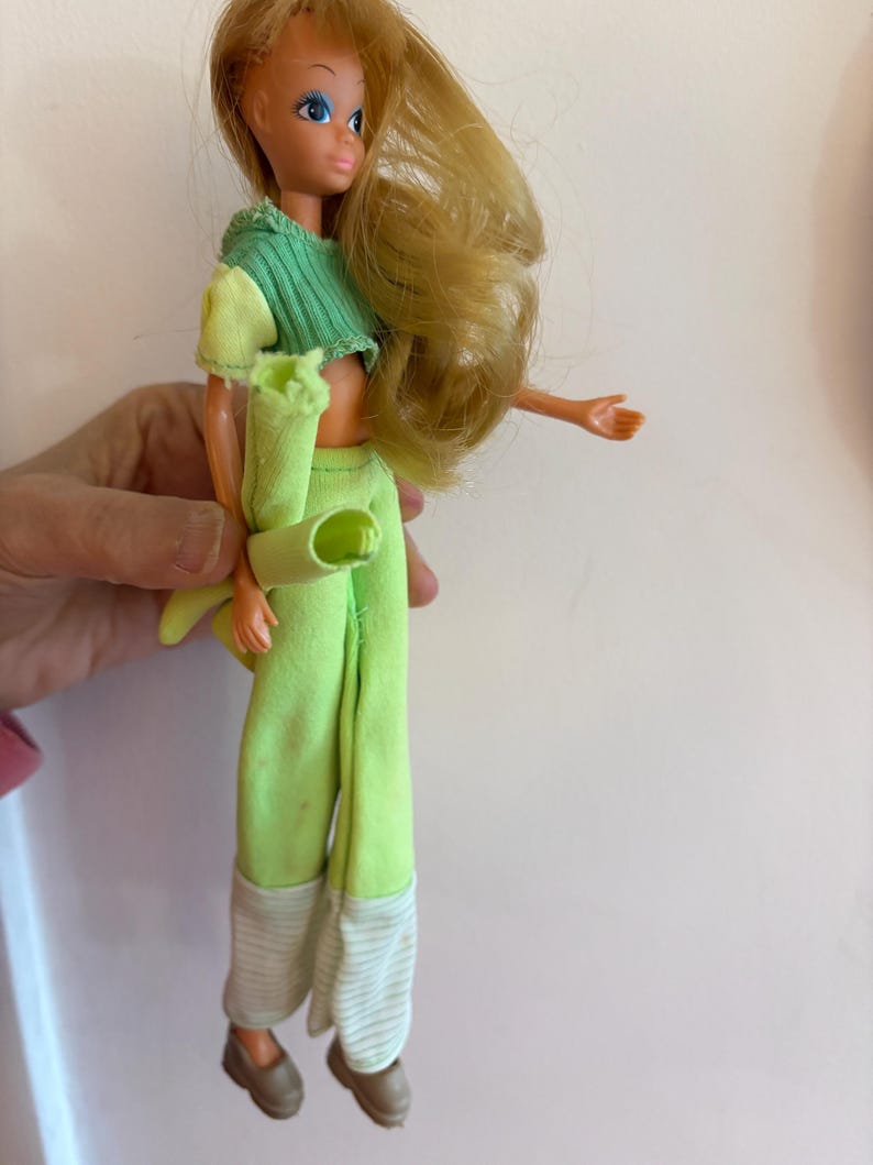 May include: A vintage doll with blonde hair and blue eyes, dressed in a green and yellow crop top, green trousers, and brown shoes. The trousers have white and grey striped cuffs. The doll is a collectible item.