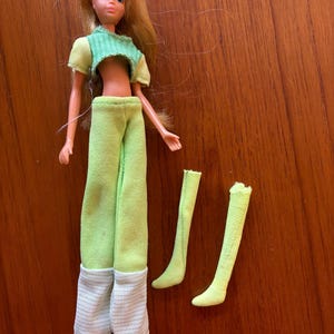 May include: A vintage doll with blonde hair, dressed in a green crop top and trousers with white and grey striped cuffs. The doll has light skin and wears white shoes. Two green socks are also included.