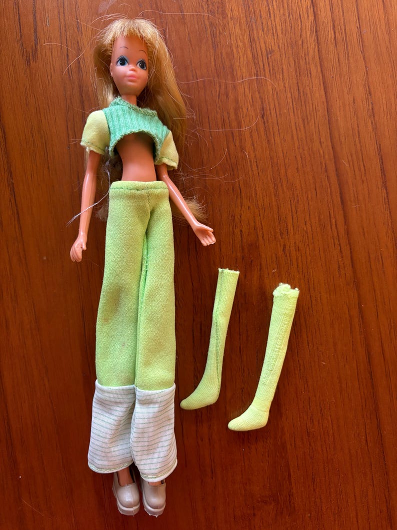 May include: A vintage doll with blonde hair, dressed in a green crop top and trousers with white and grey striped cuffs. The doll has light skin and wears white shoes. Two green socks are also included.