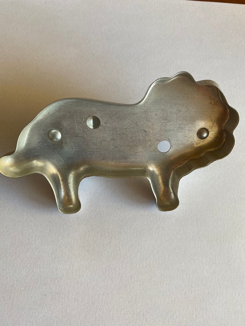 Puede incluir: Un cortador de galletas de metal plateado con forma de le&oacute;n. El cortador tiene una melena detallada y un cuerpo simple.