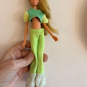 May include: A vintage doll with long blonde hair and blue eyes. The doll is wearing a green crop top, matching trousers, and brown shoes. The trousers have striped cuffs. The doll is held by a hand.