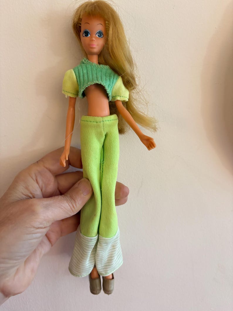 May include: A vintage doll with long blonde hair and blue eyes. The doll is wearing a green crop top, matching trousers, and brown shoes. The trousers have striped cuffs. The doll is held by a hand.