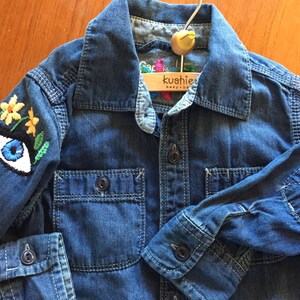 May include: A blue denim shirt with embroidered yellow flowers and a blue eye on one sleeve. The shirt has a wooden hanger with the text "kushie baby" on it.