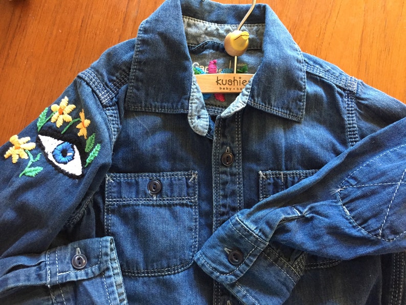 May include: A blue denim shirt with embroidered yellow flowers and a blue eye on one sleeve. The shirt has a wooden hanger with the text "kushie baby" on it.