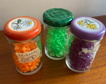 May include: Three glass jars with lids, filled with colorful beads. The jars have different colored lids and designs. The orange jar has a label that says "tri beads 107".