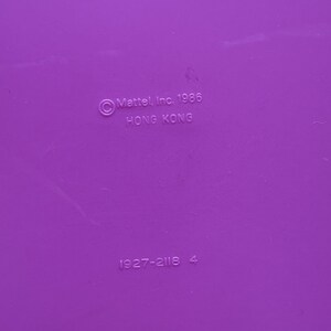 Peut inclure: Gros plan d'une surface violette unie avec le texte "&copy; Mattel, Inc. 1986 HONG KONG" et les chiffres "1927-2116 4" imprim&eacute;s dessus. La surface est lisse et uniform&eacute;ment color&eacute;e.