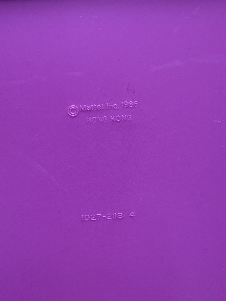 Peut inclure: Gros plan d'une surface violette unie avec le texte "&copy; Mattel, Inc. 1986 HONG KONG" et les chiffres "1927-2116 4" imprim&eacute;s dessus. La surface est lisse et uniform&eacute;ment color&eacute;e.