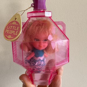 May include: A pink plastic doll in a clear plastic bottle with a purple lid. The bottle has a tag that says "Little People".