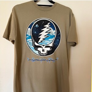 May include: Tan t-shirt featuring a circular graphic of a skull with a lightning bolt and a starry sky design. The graphic is surrounded by a blue and white border. The shirt also has text at the bottom of the graphic.