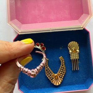 Peut inclure: Ensemble de bijoux vintage, comprenant une boucle d'oreille rose et argent&eacute;e avec des d&eacute;tails rouges, un pendentif de collier dor&eacute; et un peigne &agrave; cheveux dor&eacute;. Les articles sont dans une bo&icirc;te &agrave; bijoux rose et bleue.