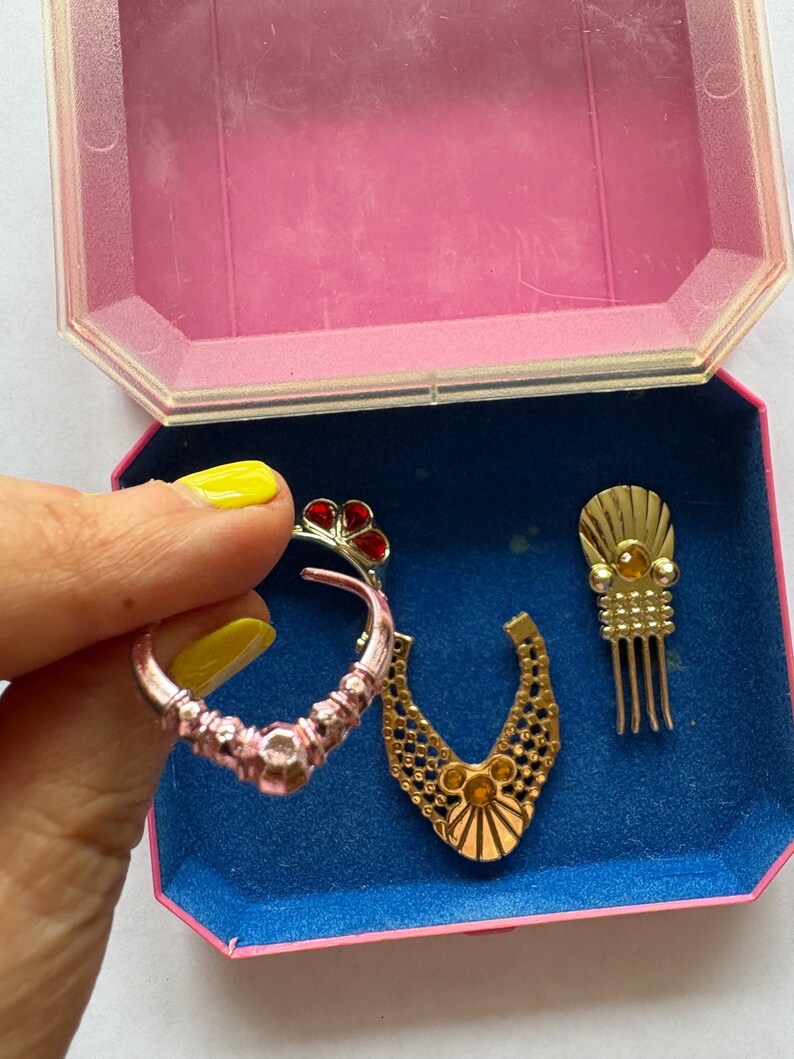 Peut inclure: Ensemble de bijoux vintage, comprenant une boucle d'oreille rose et argent&eacute;e avec des d&eacute;tails rouges, un pendentif de collier dor&eacute; et un peigne &agrave; cheveux dor&eacute;. Les articles sont dans une bo&icirc;te &agrave; bijoux rose et bleue.