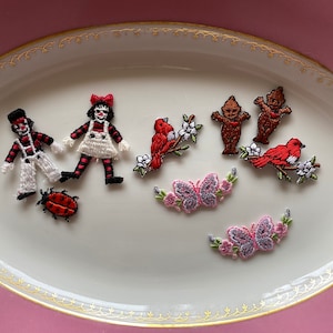 May include: A collection of embroidered iron-on patches featuring a variety of designs, including a clown, a girl in a dress, a bird on a branch, two gnomes, a ladybug, and two butterflies with pink flowers.