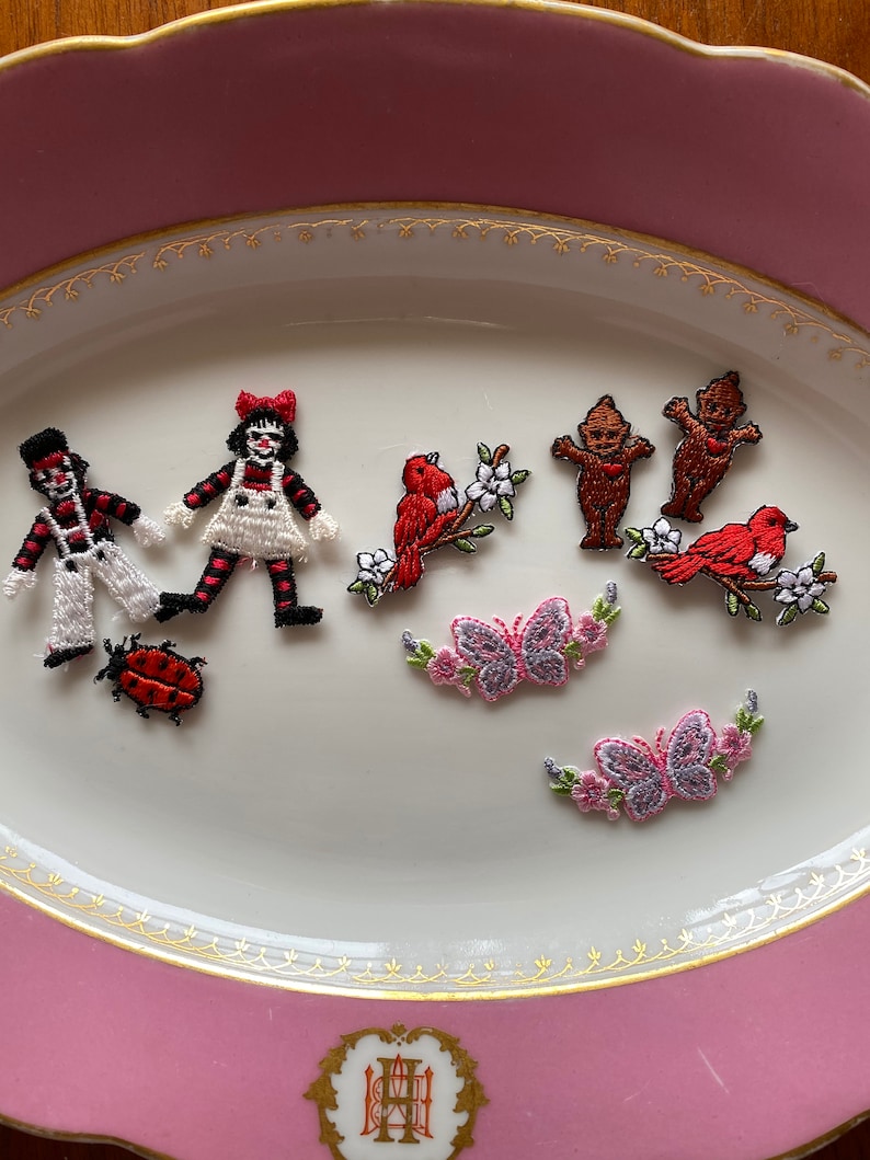 May include: A collection of embroidered iron-on patches featuring a variety of designs, including a clown, a girl in a dress, a bird on a branch, two gnomes, a ladybug, and two butterflies with pink flowers.