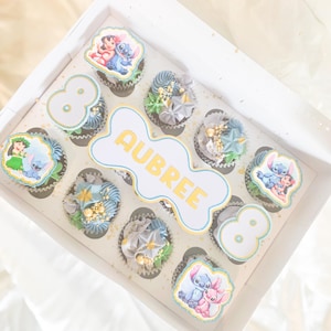 May include: A box of decorated cupcakes with a Disney theme. The cupcakes have blue, grey, and gold frosting, and some have edible images of Lilo & Stitch. The box also has the name "AUBREE" and the number "8".