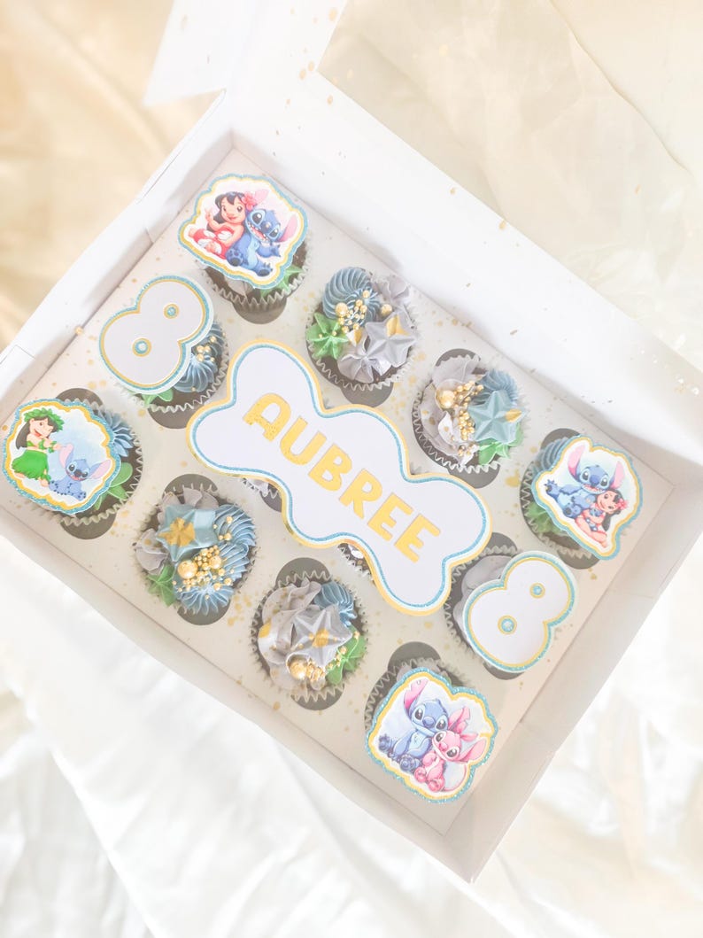May include: A box of decorated cupcakes with a Disney theme. The cupcakes have blue, grey, and gold frosting, and some have edible images of Lilo & Stitch. The box also has the name "AUBREE" and the number "8".