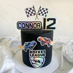 May include: A black cake decorated with Rocket League themed toppers. The cake features the name "CONNOR" and the number "12" in holographic and black glitter. Two checkered flags and car graphics complete the design.