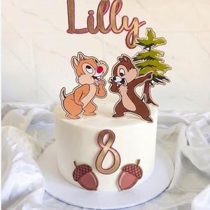 May include: A white frosted cake decorated with cartoon chipmunks, a tree, and the name "Lilly" in gold and pink glitter. The number "8" and two acorns are also featured. A festive cake for a birthday celebration.
