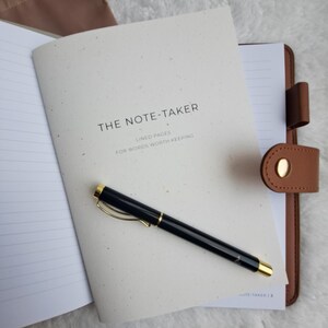 May include: A notebook with the text "THE NOTE-TAKER" and "LINED PAGES FOR WORDS WORTH KEEPING" is open, with a black pen with gold detailing resting on it. A brown leather journal with a snap closure is visible on the right.