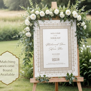 May include: A wedding welcome sign on a wooden easel, adorned with white roses and greenery. The sign features ornate gold detailing and the words "Welcome to the Nikkah" followed by names and a date. A matching welcome board is available.