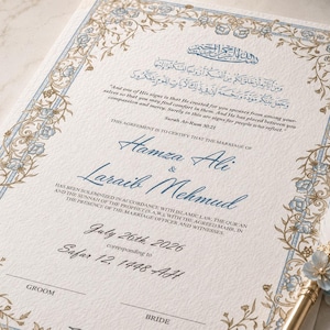 Peut inclure: Un certificat de mariage avec des bordures florales bleues et dorées, de la calligraphie arabe et du texte anglais incluant les noms Hamza Ali et Laraib Mehmud, daté du 26 juillet 2026. Une plume blanche ornée de fleurs bleues est posée à côté.