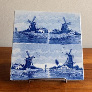 May include: A square ceramic tile featuring a blue and white transferware design. The image is divided into two horizontal panels, each depicting a landscape with windmills, small houses, a sailboat on a waterway, and birds in the sky.