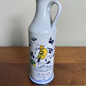 May include: A white ceramic olive oil bottle with a blue rim and handle. The bottle features a yellow bird design with blue and green accents, and the text "OLIO EXTRAVERGIN DI OLIVA".
