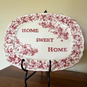 May include: An oval ceramic platter with a scalloped edge, featuring a cream-colored base and a dark red floral pattern around the border. The text "HOME SWEET HOME" is printed in dark red capital letters across the center of the platter.