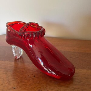 May include: A vibrant red glass shoe with a clear, block-shaped heel. The shoe features a strap detail across the top and a glossy finish. This decorative piece is a unique accent for any space.