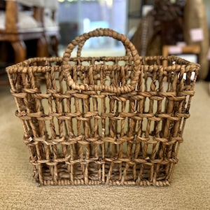 May include: A woven basket with a rectangular shape and a rounded handle. The basket is made of natural-colored, tightly woven fibers. The handle is also made of the same material. The basket is designed for carrying items.