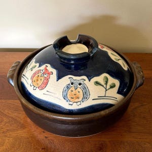May include: A round ceramic cooking pot with a dark brown base and two side handles. The lid features a deep blue top section and a cream-colored band decorated with hand-painted illustrations of two owls and small green trees.
