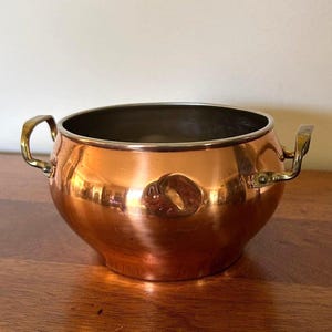 May include: A round copper bowl with a polished finish and two brass handles attached to the sides. The interior of the bowl is metallic and smooth, featuring a wide opening and a curved base that tapers toward the bottom.