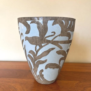 May include: A tapered ceramic vase featuring a light blue glazed surface with a visible crackle finish. The exterior is decorated with a repeating pattern of brown, stylized leafy branches and bird silhouettes wrapping around the entire vessel.